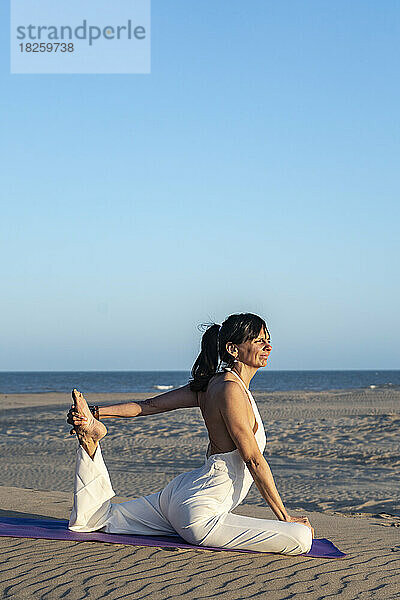 Eine Frau praktiziert Yoga auf einer Matte am Strand neben dem Meer