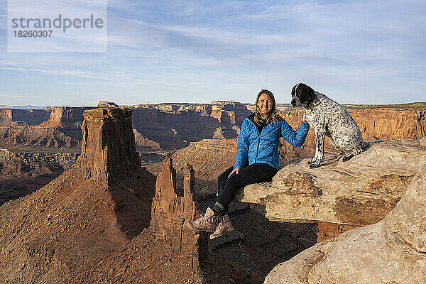 Porträt einer fröhlichen Frau  die mit Hund auf einem Felsen in der Wüste sitzt