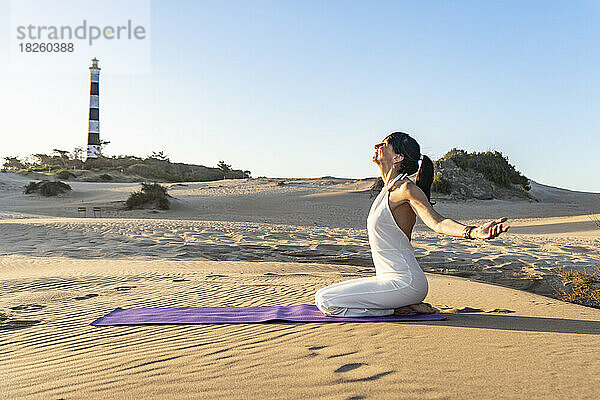 Eine Frau kniet auf einer Matte  während sie an einem sonnigen Tag in einer Düne Yoga trainiert