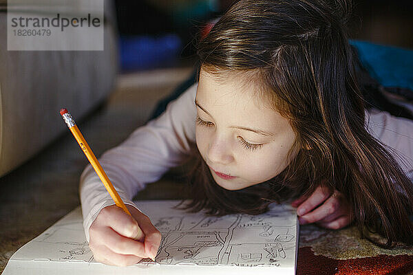 Ein kleines Mädchen liegt auf dem Boden und zeichnet sorgfältig in einen Skizzenblock