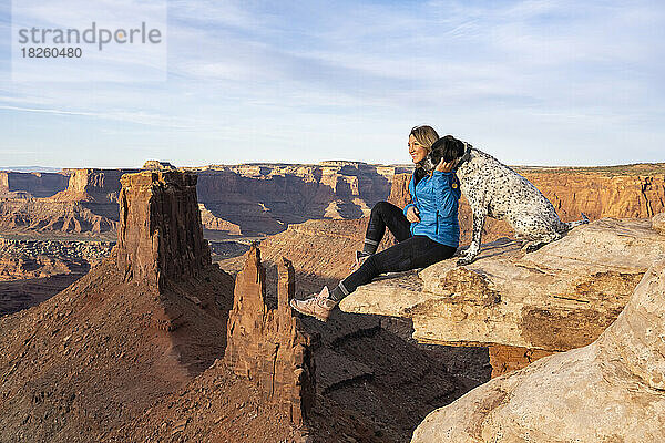Glückliche Frau blickt auf die Aussicht  während sie mit Hund auf einem Felsen in der Wüste sitzt