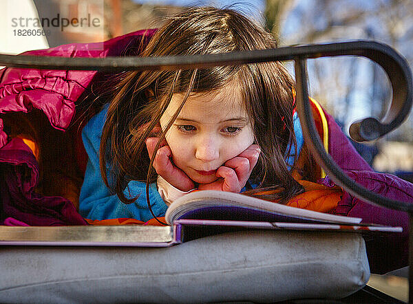 Ein kleines Mädchen legt den Kopf auf die Hände und liest draußen ein Buch