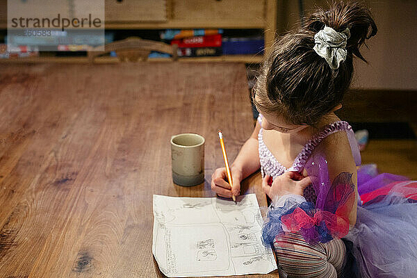 A little girl sits at table with mug drawing a comic with pencil