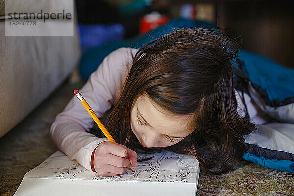 Ein kleines Mädchen  das auf dem Boden liegt  zeichnet sorgfältig im Skizzenblock