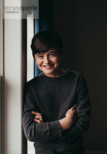 Porträt eines glücklich lächelnden Tween-Jungen  der neben einem Fenster steht.