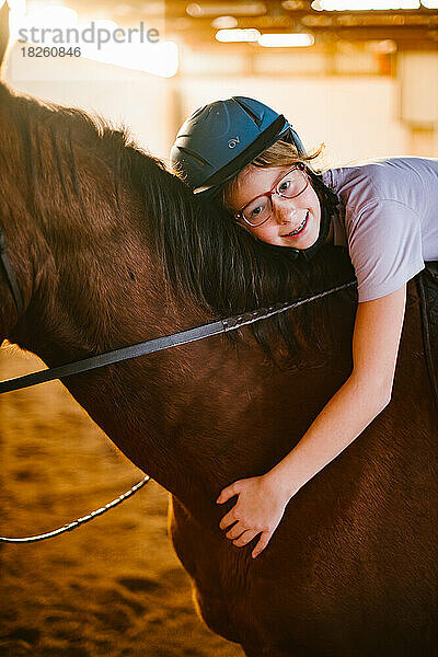 Mädchen mit Brille und Helm umarmt ihr braunes Pferd im Stall