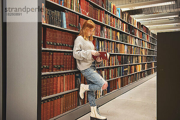 Teenager-Mädchen mit roten Haaren steht an Regalen in der Bibliothek und liest.