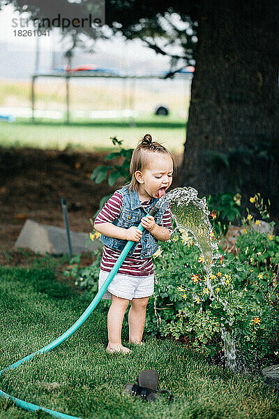 Kleines Mädchen trinkt Wasser aus einem Schlauch draußen am Garten