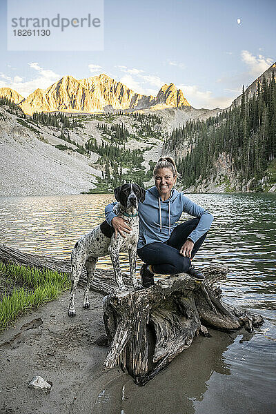 Glückliche Frau sitzt mit Hund auf Treibholz am Avalanche Lake