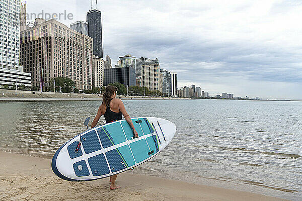 Frau trägt Paddleboard  während sie in Richtung See in der Stadt geht