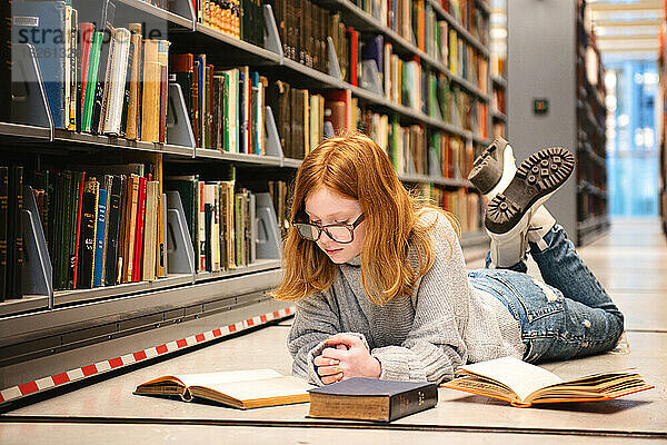 Teenager-Mädchen mit roten Haaren liegt auf dem Boden der Bibliothek und liest.