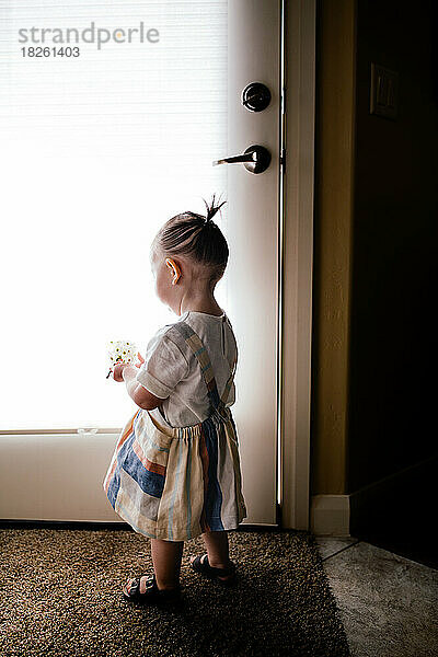 Baby trägt Kleid und hält Blumen an einem hellen Fenster im Haus