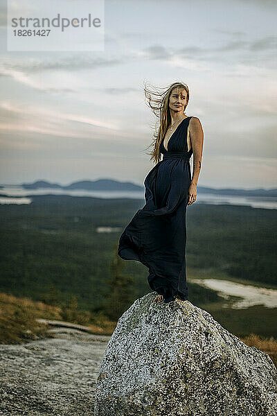 Blonde Frau im langen dunklen Kleid steht auf einem Felsen. vom Wind verweht.