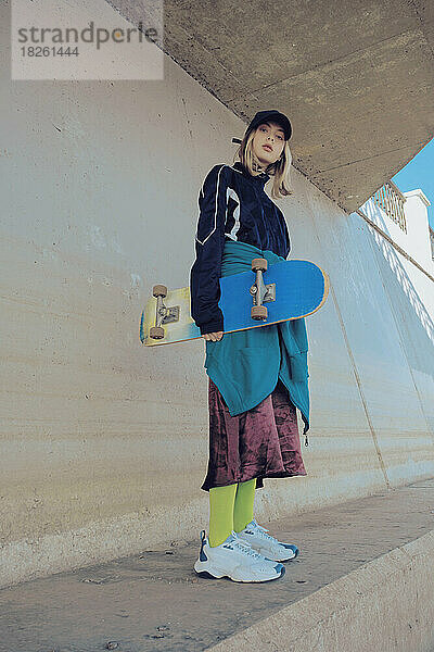Süßes Teenager-Mädchen mit Skateboard  draußen in trendiger Kleidung