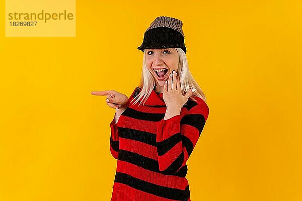 Pointing links Winter themed blonde kaukasische Mädchen auf gelben Studio Hintergrund