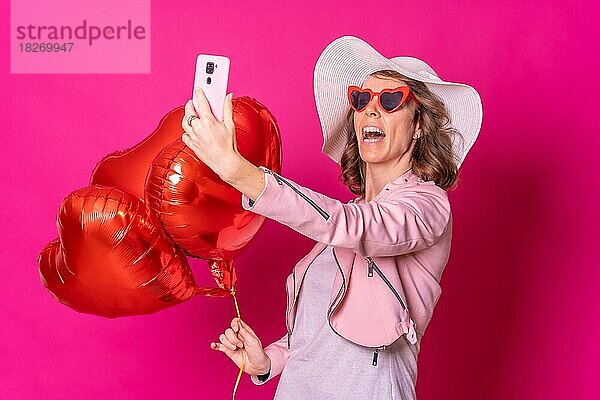 Porträt einer kaukasischen Frau  die sich mit einem weißen Hut in einem Nachtclub mit einigen Herzballons amüsiert und ein Selfie macht  rosa Hintergrund