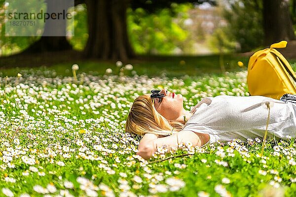 Ein junges blondes Mädchen mit Hut  das im Frühling in einem Park in der Stadt  in der Natur  auf dem Gras neben Gänseblümchen liegend  in die Kamera schaut  sich entspannt und reines Haar atmet