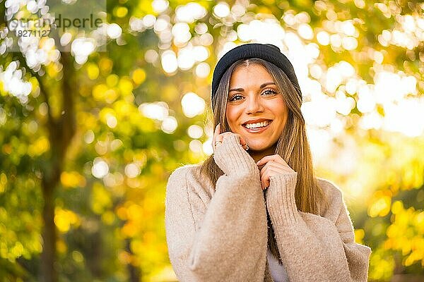 Junges blondes Mädchen mit einer Wollmütze im Herbst bei Sonnenuntergang in einem Naturpark lächelnd