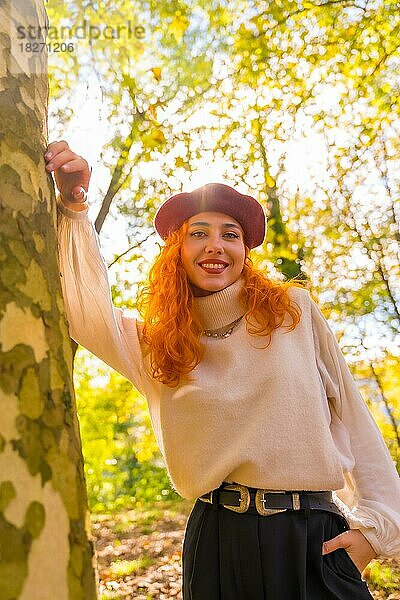 Rothaarige Frau lehnt an einem Baum in einem Waldpark und lächelt bei Sonnenuntergang