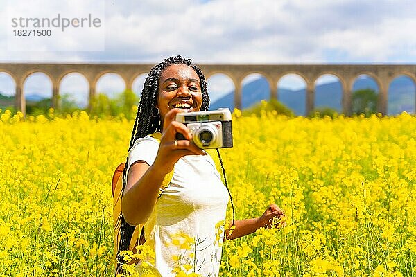 Mit einer alten Kamera lächelnd fotografieren  ein schwarzes ethnisches Mädchen mit Zöpfen  eine Reisende  in einem Feld mit gelben Blumen