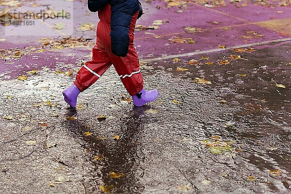 Junge in Gummistiefeln und Regenhose  der an einem regnerischen Tag durch eine Pfütze läuft