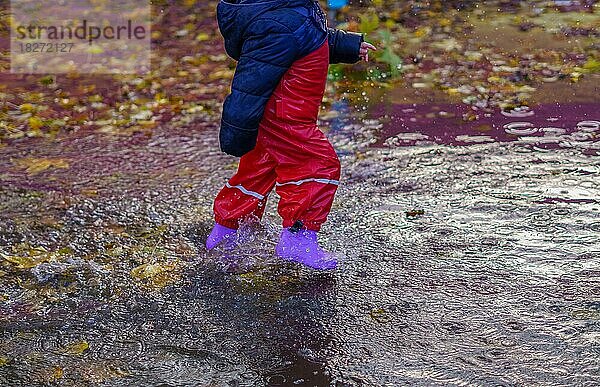 Junge in Gummistiefeln und Regenhose  der an einem regnerischen Tag durch eine Pfütze läuft