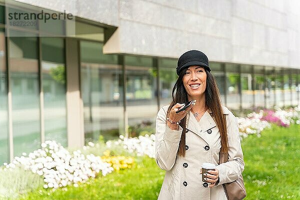 Erwachsene Geschäftsfrau vor dem Büro mit einem Kaffee in der Hand  die mit dem Telefon eine Sprachnachricht sendet