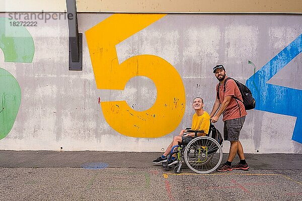 Eine behinderte Person im Rollstuhl mit einer Betonwand  die mit einem Freund spazieren geht