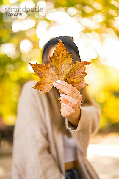 Junges blondes Mädchen mit Wollmütze im Herbst  mit einem Blatt auf dem Gesicht in den Sonnenuntergang lächelnd