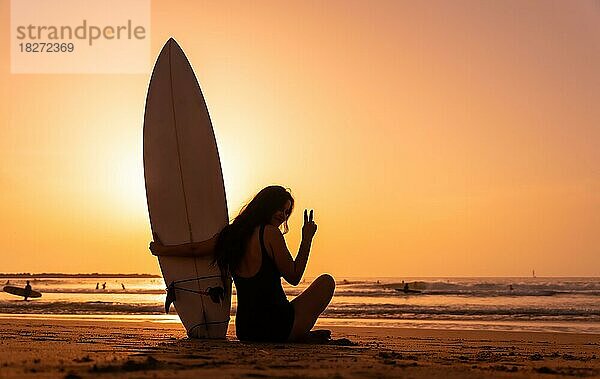 Silhouette einer Surferin am Strand bei Sonnenuntergang  die das Siegeszeichen macht