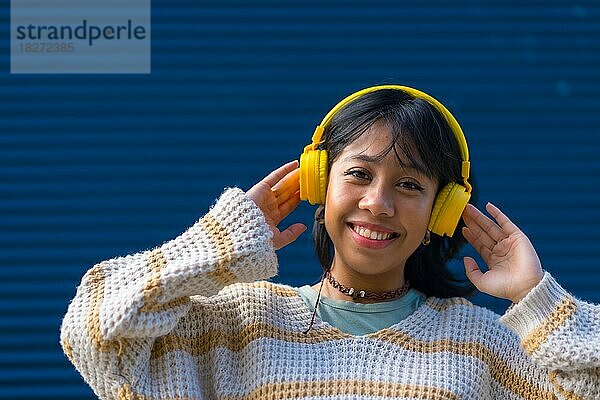 Asiatische junge Frau  die sich beim Musikhören mit gelben Kopfhörern auf einem blauen Universitätshintergrund amüsiert