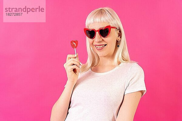 Blonde kaukasische Mädchen im Studio auf rosa Hintergrund  lächelnd Porträt in Herzen Brille mit einem Lutscher