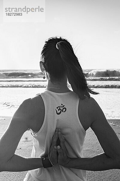 Rückansicht einer jungen Frau  die am Strand Yoga praktiziert. Om-Zeichen. Schwarz-Weiß-Aufnahme