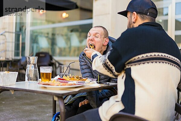 Ein Behinderter  der auf der Terrasse eines Restaurants isst  und ein Freund  der ihm beim Essen hilft
