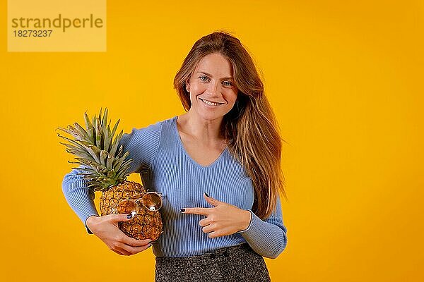 Frau mit Ananas und Sonnenbrille in einem Studio auf gelbem Hintergrund  die auf die Ananas zeigt