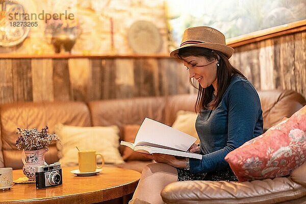 Eine hübsche Touristin mit Hut und Fotoapparat trinkt Tee in einem Café und liest ein Buch auf einem Sofa in den Räumlichkeiten