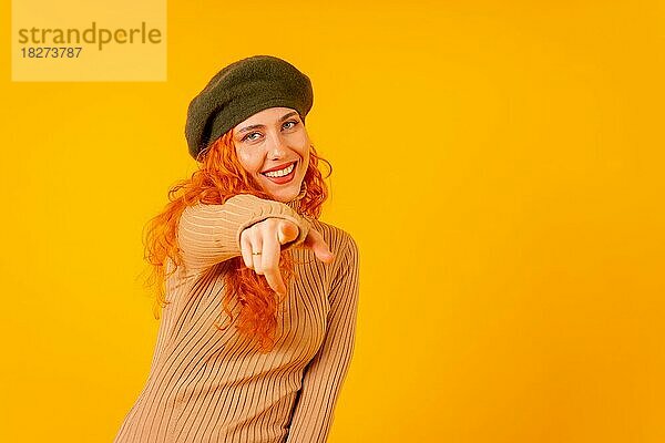 Rothaarige Frau mit Baskenmütze im Studio auf gelbem Hintergrund  Kopierraum  in die Kamera zeigend