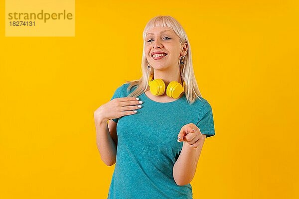 Blondes kaukasisches Mädchen  das mit Kopfhörern auf die Kamera zeigt  im Studio auf gelbem Hintergrund