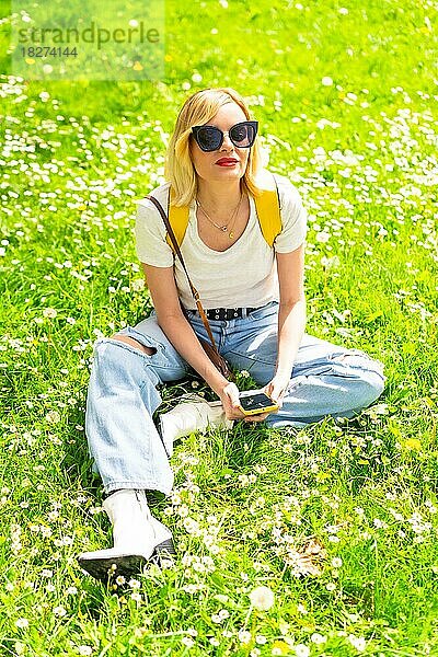 Porträt einer blonden Touristin mit Hut und Sonnenbrille  die im Frühjahr im Gras neben Gänseblümchen sitzt  in die Kamera schaut und die Natur genießt