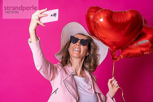 Blonde kaukasische Frau mit einem weißen Hut und einer Sonnenbrille in einem Nachtclub mit einigen Herzluftballons  die ein Selfie machen  rosa Hintergrund