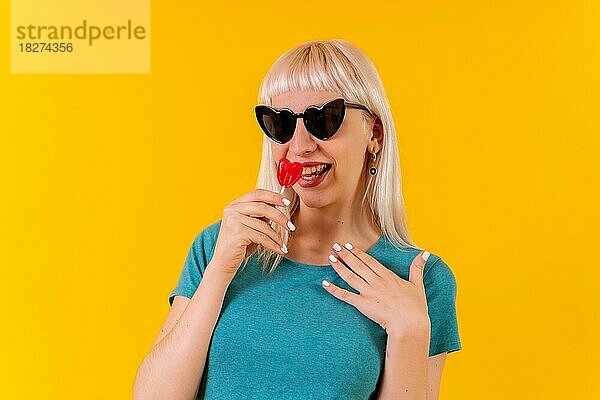 Mit Spaß essen ein Herz Lutscher  blonde kaukasische Mädchen auf gelbem Hintergrund Studio