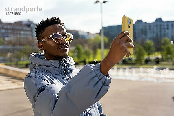 Ein schwarzer ethnischer Mann mit einem Telefon und einer blauen Jacke in der Stadt  der ein Selfie macht