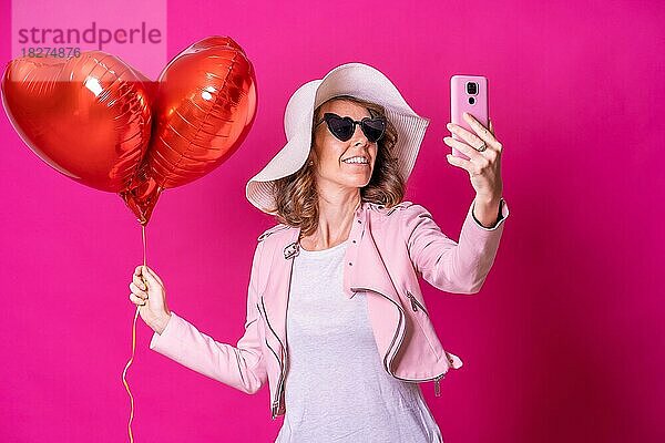 Eine kaukasische Frau  die Spaß mit einem weißen Hut in einem Nachtclub mit einigen Herz-Ballons  die ein Selfie mit ihrem Handy  rosa Hintergrund