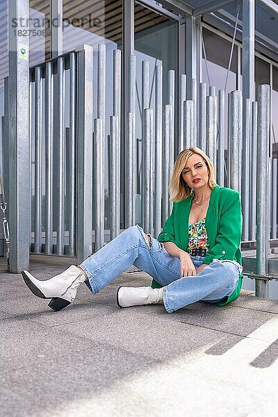 Porträt einer blonden Model-Frau in einer grünen Jacke  die auf dem Boden in einem Gebäude in der Stadt sitzt. Sommerurlaub Lebensstil