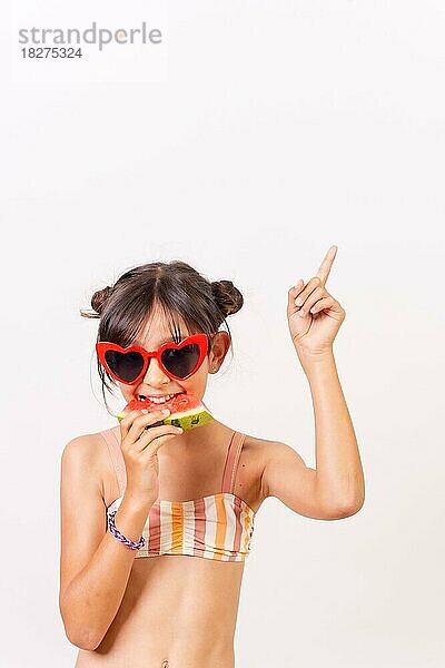 Kleines Mädchen mit Sonnenbrille isst eine Wassermelone und zeigt nach oben  Sommerferien. Weißer Hintergrund