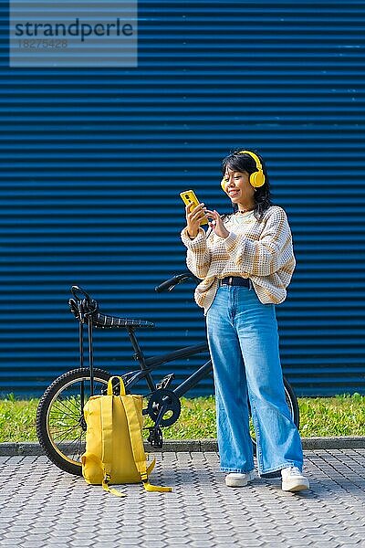 Asiatisches College-Mädchen  das mit gelben Kopfhörern auf einem blauen College-Hintergrund Musik hört