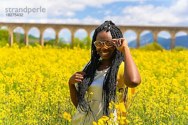 Porträt eines Mädchens schwarzer Hautfarbe mit Zöpfen und Sonnenbrille  Reisende  in einem Feld mit gelben Blumen