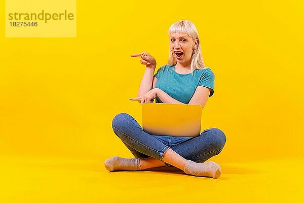 Blondes  kaukasisches Mädchen im Studio auf gelbem Hintergrund  mit einem Laptop sitzend  der nach links zeigt