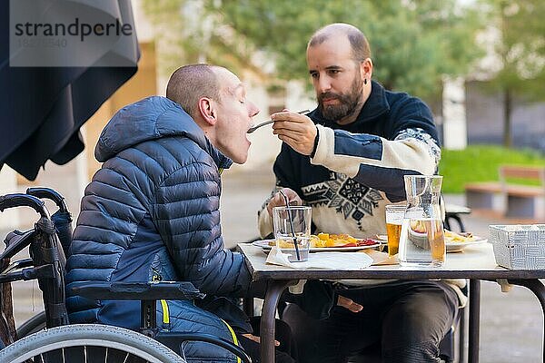 Ein Behinderter  der auf der Terrasse eines Restaurants isst  und ein Freund  der ihm beim Essen hilft