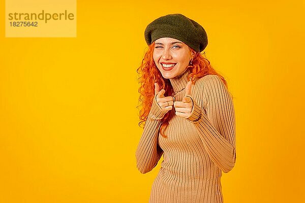 Rothaarige Frau mit Baskenmütze im Studio auf gelbem Hintergrund  Kopierraum  in die Kamera zeigend